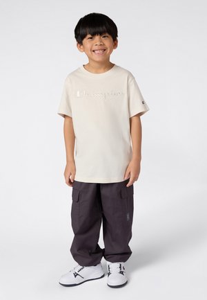 Ragazzo sorridente con capelli neri, che indossa una maglietta Champion color crema, pantaloni cargo larghi scuri e sneaker bianche, in piedi su uno sfondo neutro.