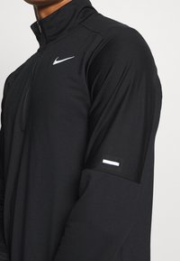 Maglia sportiva nera a mezza zip con maniche lunghe, realizzata in un tessuto testurizzato e con il logo Nike bianco sul petto. Accentuata da una striscia bianca sul braccio.