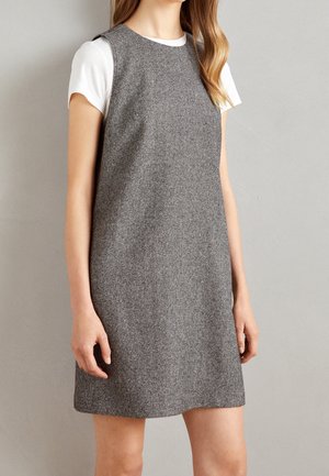 Mujer con vestido de tweed gris sin mangas sobre una camiseta blanca de manga corta, delante de una pared lisa de color gris claro.