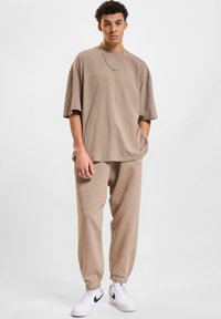 Beżowy, oversize t-shirt z krótkim rękawem, zestawiony z dopasowanymi dresami o zwężanym kroju. Sneakersy mają biały kolor z czarnymi akcentami.