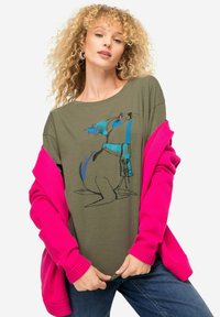 Oliwkowa koszulka z niebieskim wzorem graficznym przedstawiającym kreskowego walrusa, w zestawie z jasnoróżowym kardiganem i dżinsami.