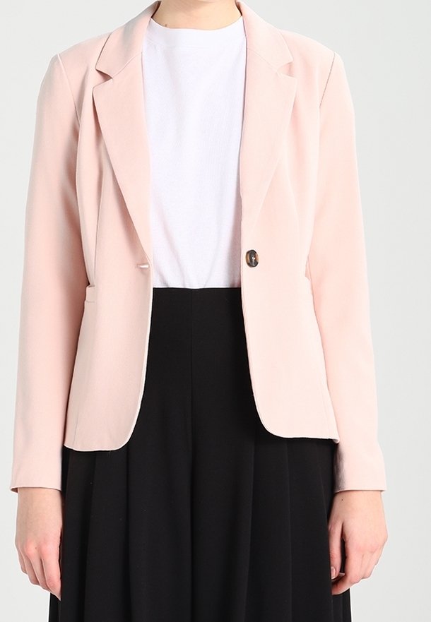 Femme portant un blazer rose clair sur un t-shirt blanc associé à une jupe noire taille haute sur un fond uni.