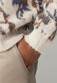 Knit sweater in cream with brown and blue abstract patterns, featuring a textured finish and ribbed cuffs. Hand in pocket of beige pants.