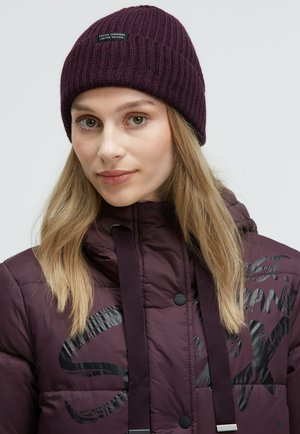 Strickmütze in Lila mit geripptem Muster und einem Logo-Patch, getragen mit einer dunkelvioletten Daunenjacke, die einen grafischen Druck und Kordelzüge hat.