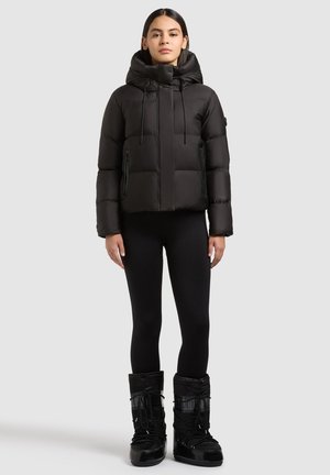 Femme portant une doudoune noire avec capuche, des leggings noirs et des bottes d'hiver noires, debout devant un fond clair uni.