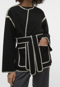 Schwarze Jacke mit gewelltem weißen Saum, langen Ärmeln, zwei Taschen und einem selbstgebundenen Gürtel. Hergestellt aus strukturiertem Stoff, der eine strukturierte Passform bietet.
