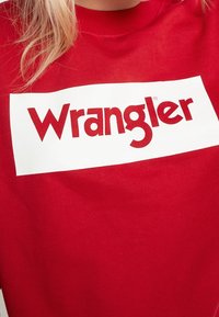 Camiseta de algodón roja con un parche rectangular blanco que muestra el texto "Wrangler" en una fuente roja en negrita. El diseño es simple y gráfico.