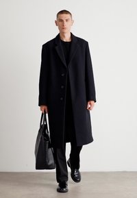 Abrigo de lana negro con cuello de muesca y cinco botones, combinado con pantalones y zapatos negros a juego, y un bolso tote negro.