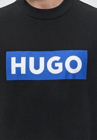 Schwarzes Crewneck-Shirt mit einem blauen rechteckigen Logo in der Mitte der Brust, das den weißen Schriftzug „HUGO“ zeigt.