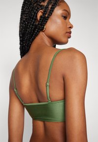 Grønn sateng-bralette med tynne stropper, som viser en jevn tekstur og subtil glans, med justerbare kroker på baksiden for en sikker passform.