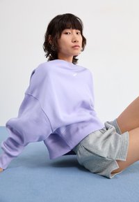 Light purple oversized sweatshirt with ribbed cuffs and hem, featuring a small white logo. Paired with gray shorts on a blue surface.