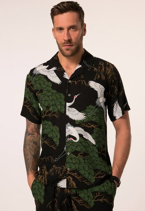 Mann mit Bart, der ein schwarzes kurzärmeliges Hemd mit Knöpfen und Hose mit einem Muster aus weißen Kranichen und grünen Bäumen trägt, steht mit den Händen in den Taschen.