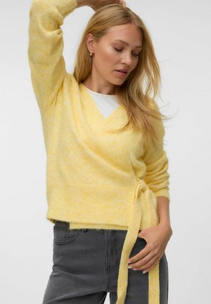 Femme aux longs cheveux blonds portant un pull cache-cœur jaune doux et un jean gris foncé, posant avec un bras levé et les yeux fermés.