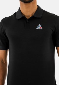 Camiseta polo negra hecha de un tejido texturizado, con cuello, mangas cortas y un logotipo bordado en azul, rojo y blanco en el pecho.
