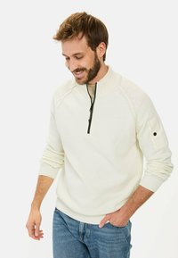 Cremefarbener Strickpullover mit hohem Kragen, halbem Reißverschluss, gerippten Bündchen und einem dezenten strukturierten Muster an den Ärmeln. Kombiniert mit blauen Jeans.