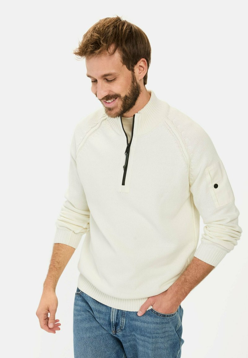 Cremefarbener Strickpullover mit hohem Kragen, halbem Reißverschluss, gerippten Bündchen und einem dezenten strukturierten Muster an den Ärmeln. Kombiniert mit blauen Jeans.