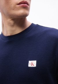 Άνδρας που φοράει ένα υφασμάτινο μπλε ναυτικό πουλόβερ Calvin Klein με ένα μικρό κόκκινο και λευκ ό έμβλημα CK στο αριστερό στήθος, κοιτάζοντας προς τα πλάγια.