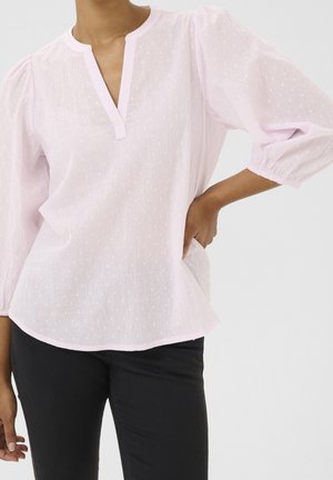 Kvinde iført lyserød tekstureret bluse med trekvartlange pufærmer og sorte bukser, hånd hvilende på hoften mod en ensfarvet baggrund.