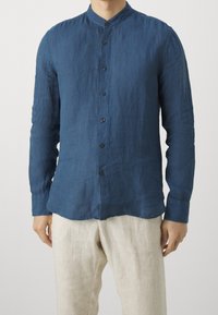 Hombre vestido con una camisa de manga larga azul marino abotonada y un collar mao, y pantalones de lino beige, contra un fondo liso.