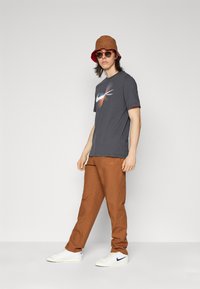 Gray Nike t-shirt with colorful logo, brown pants, and white sneakers. Accessories include a brown bucket hat and sunglasses.
