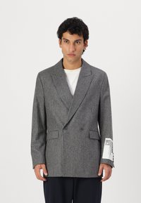 Blazer gri cu model houndstooth, cu design cu două rânduri de nasturi, revere late, buzunare frontale și detaliu cu etichetă pe mânecă. Material neted, texturat.