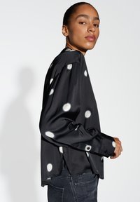 Black satin long-sleeve top featuring large white dots, a relaxed fit, side slits, and buttoned cuffs. Smooth texture and subtle sheen.