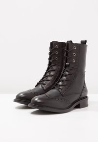Stivali alla caviglia in pelle nera con un dettagliato design brogue, chiusura con lacci, punta rotonda e tacco basso a blocco. Texture liscia con finitura elegante.