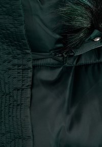 Tessuto di giacca trapuntata verde scuro con una superficie liscia, dotata di un profilo in pelliccia sintetica removibile e un dettaglio di chiusura a scatto.