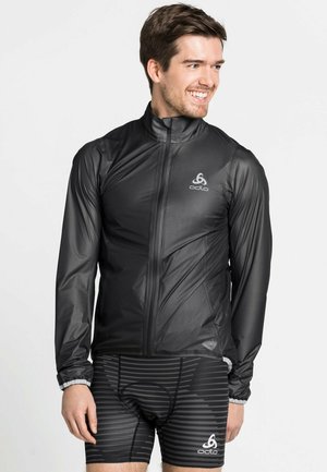 Leichte schwarze Sportjacke mit hohem Kragen, durchgehendem Reißverschluss und reflektierendem Logo. Getragen über gestreiften schwarzen Shorts.