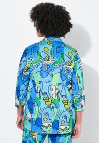 Ce haut en polyester léger et coloré présente un design abstrait en bleu, vert et beige avec des visages et des illustrations au trait. Manches trois-quarts.