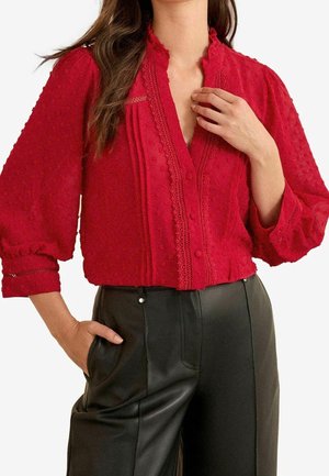 Kvinde iført en rød tekstureret bluse med blonde detaljer og sorte læderbukser med høj talje, hånd i lomme og fingre nær kraven.