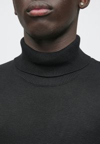 Pull noir à col roulé avec texture côtelée et col à double couche. Vue en gros plan centrée sur la zone du cou, mettant en avant un tissu lisse et un design simple.