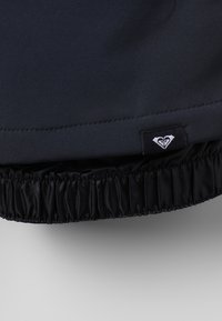 Sort jakke med en glat tekstureret overflade og elastisk talje, med et lille logo-tag i hvid.