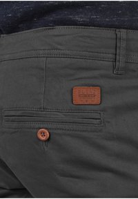 Pantaloni grigi scuri con un tessuto testurizzato. Presentano un'etichetta in pelle con la scritta "BLEND", una chiusura con bottone e una tasca posteriore con dettagli di cucitura.
