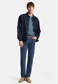 Camicia in denim scuro con bottoni, maglione teal, pantaloni blu, stivali beige, slim fit, cuciture minime e una tasca sul petto della camicia.