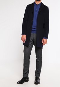 Manteau en laine bleu marine sur un polo bleu, associé à un pantalon gris à motifs et des chaussures en cuir noires. Coupe ajustée, design boutonné, texture lisse.