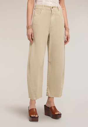 Kvinde iført beige bredbenede korte bukser og brune platformsandaler, stående med hænder afslappet langs siderne.