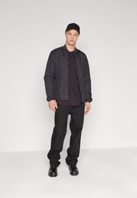 Giacca nera trapuntata con zip frontal, indossata sopra una t-shirt nera, abbinata a pantaloni neri larghi e scarpe nere, su uno sfondo neutro.