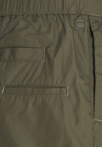 Tissu vert olive avec des ouvertures de poche rectangulaires cousues et un petit logo en caoutchouc surélevé portant l'inscription "RAW" près de la taille.