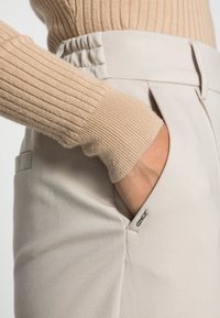 Beiger, gerippter Strickpulloverärmeln, dazu hellgraue Hose mit seitlicher Tasche und dezentem Markenakzent. Weicher, strukturierten Stoff.