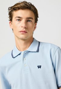 Ljusblå piké i texturerad tyg, med tvåknappsstängning, marinrandig krage och en liten marinblå logotyp på bröstet.