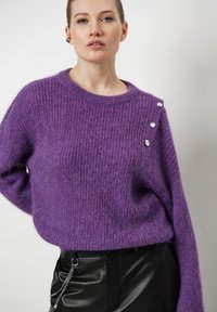 Paarse gebreide trui met een ronde hals, losse pasvorm en drie decoratieve knopen op de schouder; gecombineerd met zwarte leren broeken.