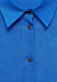 Dettaglio ravvicinato di una camicia blu con colletto e tre bottoni marroni visibili lungo il davanti.
