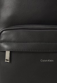 Sac à dos en cuir noir avec une finition texturée, poche avant avec quincaillerie en métal, et logo "Calvin Klein" embossé sur le côté.
