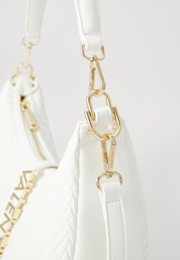 White leather handbag featuring a quilted texture, gold hardware, and a prominent brand logo. Includes a detachable strap and zipper detail.