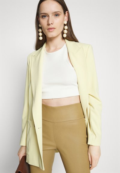 Ανοιχτό κίτρινο κομμένο σακάκι πάνω από λευκό crop top και ταλαντευμένα παντελόνια σε καφετί απόχρωση, με μεγάλα κρεμ χρωματιστά κόσμημα για τα αυτιά.
