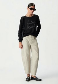 Zwarte trui met tekst "Feeling Rock", lichtbeige wijde broek, zwarte loafers en zonnebril. Gestructureerde brei en ontspannen pasvorm.