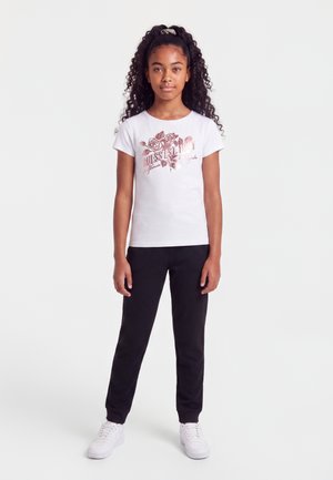 Pige med krøllet hår står iført en hvid t-shirt med blomstermotiv, sorte bukser og hvide sneakers mod en ensfarvet hvid baggrund.