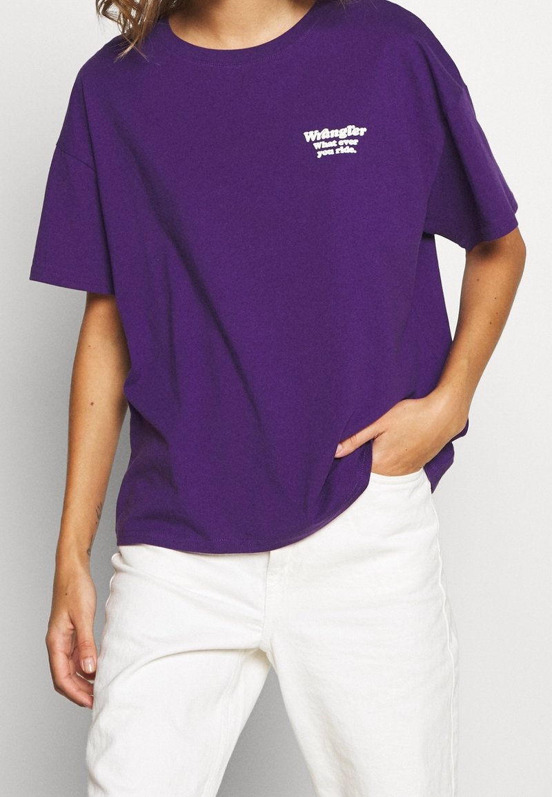 Lila bomullströja med korta ärmar och rund hals. Har en vit textlogotyp på vänster bröst. Stylingen inkluderar vita byxor.