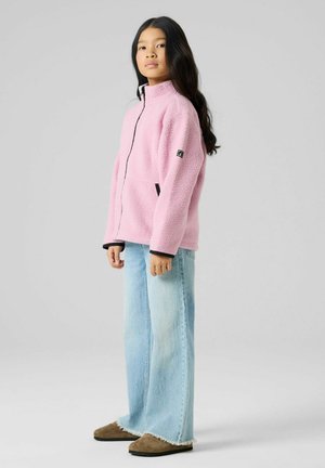 Mladi človek stoji v stranski drži, oblečen v svetlo roza flis jakno, svetlo modre široke hlače in rjave natikače.